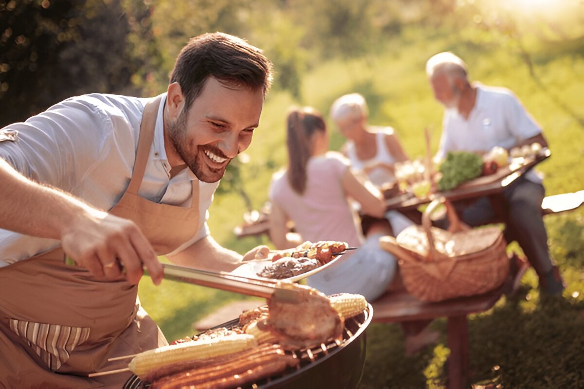 Mid-Thirties Man Using BBQ Tools and Smoker Accessories to Prepare Lunch on Summer Afternoon with Senior Mother, Father, and Wife